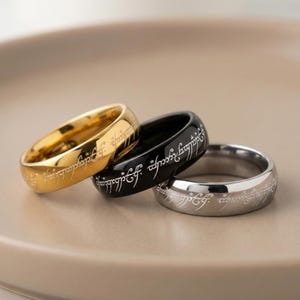 K&ouml;nnte beinhalten: Drei Ringe in Gold, Schwarz und Silber, jeweils mit eingravierter Schrift. Der goldene Ring befindet sich links, der schwarze Ring in der Mitte und der silberne Ring rechts. Die Ringe liegen auf einer neutral get&ouml;nten Oberfl&auml;che.