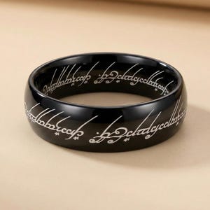 K&ouml;nnte beinhalten: Ein schwarzer Ring mit wei&szlig;er, eingravierter Schrift um das Band. Der Ring hat eine polierte, reflektierende Oberfl&auml;che. Die Schrift scheint in einer stilisierten Fantasiesprache zu sein. Der Ring ist ein massives Band mit glattem, abgerundetem Rand.