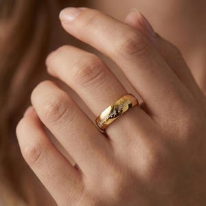 K&ouml;nnte beinhalten: Ein goldfarbener Ring mit eingravierter Schrift. Der Ring wird an einem Finger getragen. Die Schrift ist in einer stilisierten Schriftart. Der Ring ist im Bandstil. Der Ring besteht aus gl&auml;nzendem Metall.
