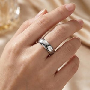 K&ouml;nnte beinhalten: Ein silberner Ring mit eingraviertem Text. Der Ring wird an einem Finger getragen und scheint aus einem gl&auml;nzenden Metall zu bestehen. Der Text ist klein und detailliert und umschlie&szlig;t das Band des Rings.