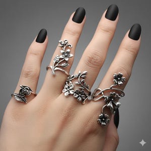 May include: A hand displays five silver-toned floral rings. Each ring features a different design of flowers and leaves. The rings are worn on various fingers, showcasing the intricate details of the botanical-inspired jewelry. The background is a solid gray.