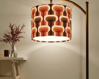 May include: A retro-style floor lamp with a cylindrical lampshade. The shade features a repeating pattern of brown, orange, and cream-colored shapes. The lamp has a curved, gold-toned arm and a dark brown pole. A small table with a vase of red flowers is in the background.