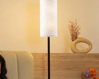 May include: A tall, black floor lamp with a cylindrical, off-white linen shade. The lamp is positioned in front of a light-colored wall and a wooden headboard. A decorative vase with dried flowers sits on the headboard.