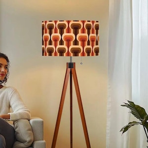 May include: A retro-style floor lamp with a wooden tripod base and a rectangular lampshade. The lampshade features a repeating pattern in shades of brown, orange, and cream. The lamp is positioned in a living room setting.