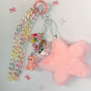 Peut inclure: Un porte-clés aux couleurs pastel avec un charm en forme d'étoile rose, un petit ours en peluche rose et une chaîne de maillons irisés. Le porte-clés comporte également un fermoir en forme d'étoile rose et plusieurs petits nœuds décoratifs.