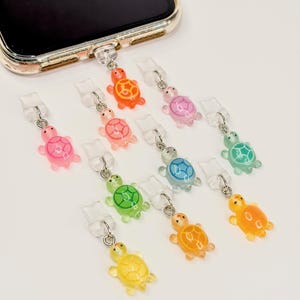 Può includere: Una collezione di ciondoli per telefono a forma di tartaruga colorati. I ciondoli sono in vari colori, tra cui rosa, arancione, giallo, verde e blu. Ogni ciondolo ha un anello di metallo color argento e un connettore di plastica trasparente.