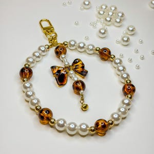 Peut inclure: Un porte-clés perlé avec un mélange de fausses perles blanches, de perles à motif écaille de tortue et d'accents dorés. Un charm en forme de nœud papillon écaille de tortue et un fermoir doré complètent le design. Des perles éparpillées ajoutent une touche décorative.