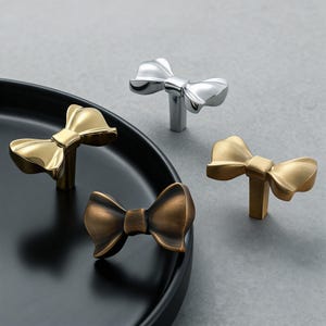 May include: Four bow-shaped cabinet knobs in gold, silver, and bronze tones. The knobs are arranged on a black plate and a gray surface. The bow details are prominent, and the knobs have a modern, decorative design.