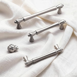 May include: A collection of brushed nickel cabinet hardware, including two long pulls, one short pull, and a knob. The hardware features a fluted design on the ends and a smooth, cylindrical bar. The items are displayed on a white, textured fabric.
