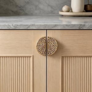 May include: Close-up of a light wood cabinet with two doors. Each door has a half-circle, textured gold handle. The cabinet doors have vertical, ridged detailing. A marble countertop is visible above the cabinet.
