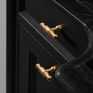 Könnte beinhalten: Nahaufnahme von zwei goldfarbenen Schrankgriffen mit T-Bar-Design. Die Griffe sind an einer schwarzen Schranktür montiert. Die Schranktür ist Teil eines größeren schwarzen Schranks mit einer schwarzen Marmorarbeitsplatte.