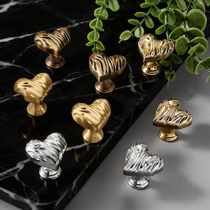 May include: A collection of heart-shaped cabinet knobs in various metallic finishes, including gold and silver. The knobs feature a textured, ridged design and are mounted on short, decorative bases. Decorative hardware.