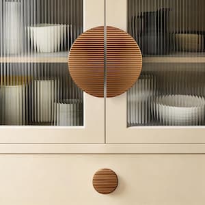 May include: A cream-coloured cabinet with ribbed glass doors and bronze circular handles. The cabinet doors are closed, revealing shelves with white and beige ceramic items. A smaller, matching circular knob is on the drawer below.