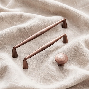 Op de afbeelding: Twee langwerpige koperkleurige metalen ladehandgrepen en een bijpassende ronde kastknop. Elke handgreep is gegraveerd met de tekst Dailey & Abbey. De ronde knop heeft een gegraveerde decoratieve rand met de tekst Est. 2020 en K & Q.
