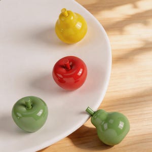 May include: A collection of colorful, fruit-shaped decorative objects. The objects include a yellow pear, a red apple, a green apple, and a green pear. The objects are glossy and appear to be made of ceramic or a similar material.