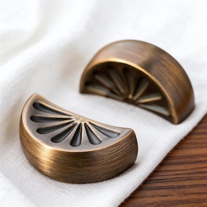 May include: Two bronze-coloured cabinet pulls shaped like citrus fruit slices. Each pull has a semi-circular shape with a detailed fan-like design on the interior. The pulls are displayed on a white cloth, suggesting a home decor or hardware theme.