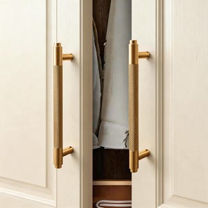 May include: Two gold-coloured cabinet handles with a textured, diamond-patterned grip. The handles are mounted on a white cabinet door. The cabinet door is slightly ajar, revealing the interior of the cabinet.