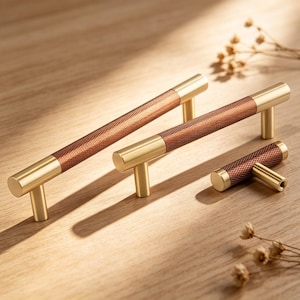 May include: Three cabinet pulls with a gold finish and brown textured centre sections. The pulls are arranged on a light wood surface, with a small sprig of dried flowers in the background. The hardware is designed for drawers and cabinets.