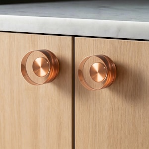 May include: Close-up of two copper-colored cabinet knobs. Each knob has a circular design with a central disc and a surrounding ring. The knobs are mounted on light-colored wooden cabinet doors, with a marble countertop visible above.