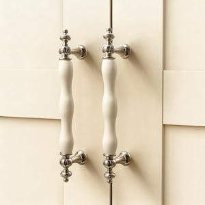 Peut inclure: Deux poignées de placard blanches et argentées au design classique. Les poignées ont une poignée blanche incurvée et allongée et une quincaillerie de montage argentée ornée. Elles sont montées sur une porte d'armoire crème.