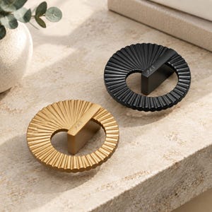May include: Two circular cabinet knobs, one in matte gold and one in matte black, featuring a radial fluted design and a central bar handle. Each knob is engraved with the letters P&S on the top surface of the bar, showcasing a textured finish.