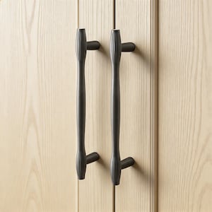 Op de afbeelding: Twee donker bronzen kastgrepen met verticale geribbelde details. De grepen zijn gemonteerd op een lichte houten kastdeur. De grepen hebben een lang, slank ontwerp met een gebogen handvat en een kleine, ronde basis.