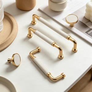 May include: Cabinet hardware in gold with white leather-effect grips. The image shows several handles and knobs. The handles are curved, and the knobs have a circular white centre. The hardware is displayed on a white surface.