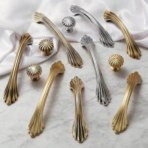 May include: An assortment of decorative cabinet hardware, including handles and knobs. The handles are in gold and silver finishes, featuring a fluted design. The knobs have a flower-like pattern.