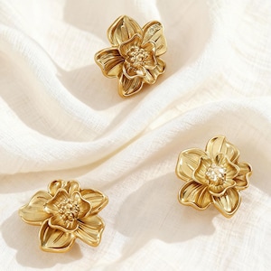 Può includere: Tre pomelli dorati a forma di fiore con dettagliati disegni a petalo. I pomelli sono disposti su un tessuto bianco strutturato, mettendo in mostra i loro intricati motivi floreali. Un pomello ha un centro di cristallo.