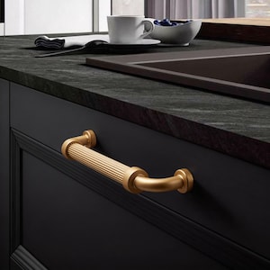 Può includere: Una maniglia dorata per armadio con un centro scanalato e estremità curve è montata su una porta di un armadio nero. L'hardware ha una calda tonalità metallica, in contrasto con i mobili scuri. L'ambiente della cucina comprende un piano di lavoro, un lavandino e una tazza.