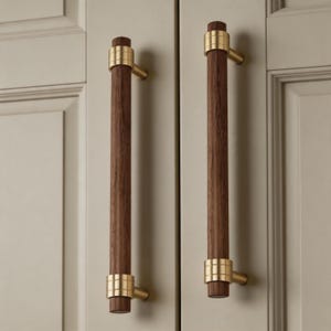 May include: Two cabinet pulls with a dark brown, cylindrical wooden handle and gold-colored metal accents. The pulls are mounted on a light beige cabinet door. The hardware has a modern, minimalist design.