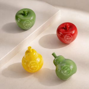 May include: Two glossy ceramic apples in green and red, and two glossy ceramic pears in yellow and green. Each fruit features a white printed emblem with the text Est. 2020 K & Q and a small ring icon, centered on the front of the smooth surface.