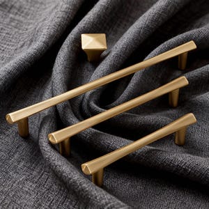 Op de afbeelding: Een verzameling goudkleurige kastbeslag, waaronder drie stanggrepen van verschillende lengtes en een vierkante knop. Het beslag wordt gepresenteerd op een grijze, getextureerde stof, wat het moderne ontwerp benadrukt.