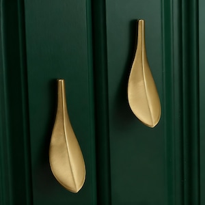 May include: Two gold-coloured leaf-shaped cabinet pulls against a dark green cabinet door. The pulls have a smooth, matte finish and are mounted vertically. The cabinet door has vertical grooves.