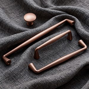 Puede incluir: Un conjunto de herrajes para armarios de color cobre, que incluye dos tiradores, un tirador más pequeño y un pomo redondo. Los herrajes se muestran sobre una tela gris texturizada, mostrando el acabado metálico y el diseño.