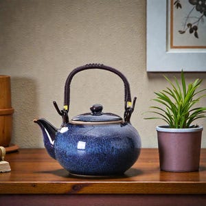 Könnte beinhalten: Eine blaue Keramik-Teekanne mit einem geflochtenen Griff und einem kleinen Deckel. Die Teekanne hat eine dunkelblaue Glasur mit helleren blauen Streifen. Der Griff ist dunkelbraun und der Ausguss ist schwarz. Die Teekanne steht auf einer Holzoberfläche.