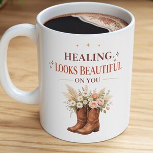 May include: White ceramic mug with the text "HEALING LOOKS BEAUTIFUL ON YOU" in brown. The mug features a floral and cowboy boot design. The mug is filled with a dark liquid and has a white handle.