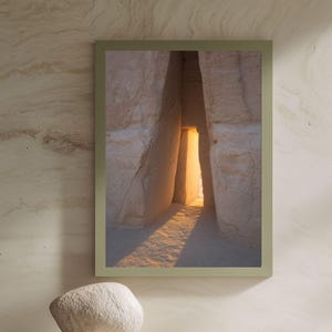 Könnte beinhalten: Ein gerahmter vertikaler Kunstdruck, der eine schmale, vertikale Öffnung zwischen zwei großen, strukturierten beigen Felsformationen zeigt. Helles, warmes goldenes Licht scheint durch den Spalt und wirft einen langen Schatten auf den Sand.