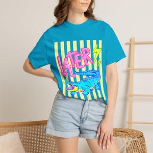May include: A turquoise t-shirt featuring a graphic design with the word "LATER" in pink, a blue alligator, and yellow lightning bolts. The shirt has a crew neck and short sleeves. The design is set against a background of yellow and white vertical stripes.