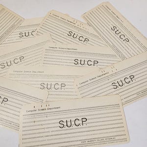 May include: A collection of vintage computer punch cards. The cards are off-white with rows of punched holes and printed text, including "Computer Science Department" and "S.U.C.P.". The cards are scattered across a white surface.