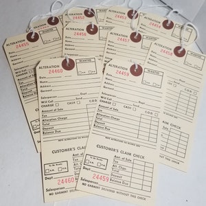 May include: Nine vintage white paper tags with black text and red and brown circular labels. The tags are for alterations and have the text "ALTERATION" and a number from 24452 to 24460. The tags have fields for date, name, address, garment, salesperson, will call, charge, amount of sale, tax, alteration charge, total, deposit, and balance due. The tags also have a "WANTED" box with checkboxes for AM and PM. The tags have a "CUSTOMER'S CLAIM CHECK" section with fields for amount of sale, tax, alteration charge, total, deposit, and balance due. The tags have a "NO GARMENT DELIVERED WITHOUT THIS CHECK" message.
