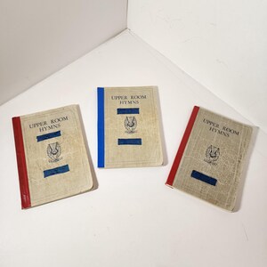 May include: Three vintage "Upper Room Hymns" books with aged, off-white covers. Each book has a different colored spine: red or blue. The covers feature blue rectangles and a crest. The books are arranged on a white surface.
