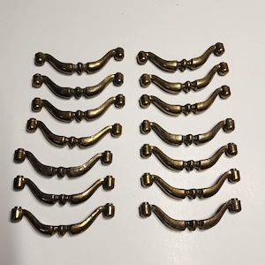 May include: Twelve antique brass cabinet pulls with a curved design. The pulls have a decorative detail in the center.