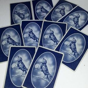May include: A collection of playing cards with a dark blue background and a white oval design. Each card features a detailed illustration of a crow holding playing cards, perched on a branch. The cards are arranged in a scattered pattern.