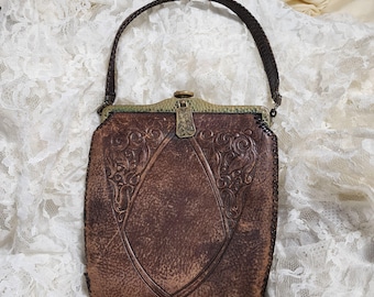 May include: A vintage brown leather handbag with an ornate embossed design. The bag features a braided handle, a decorative clasp, and a stitched border. The central design is a diamond shape with floral scrollwork.