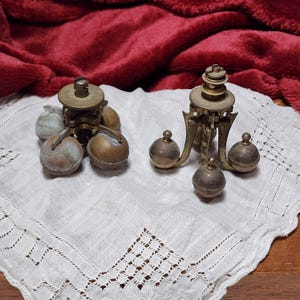 May include: Two vintage brass clock pendulum governors featuring three spherical weights attached to a central gear mechanism. The metal components show signs of age, including oxidation and patina, with intricate structural details.