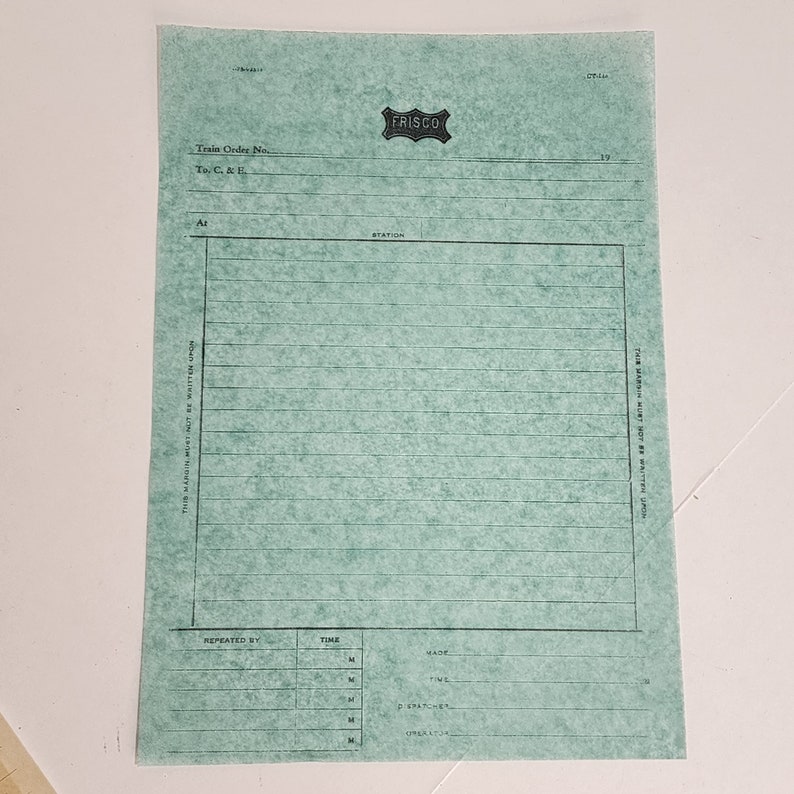 May include: A vintage teal-colored Frisco train order form. The form features a large rectangular space for writing, with horizontal lines and text including "Train Order No." and "Station." The Frisco logo is at the top.
