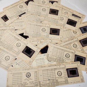 May include: A collection of vintage dual-purpose engineering document cards. The cards are beige with black text and a small rectangular window. Each card has a circular emblem and numerous rows of numbers and symbols. The cards are scattered across a surface.