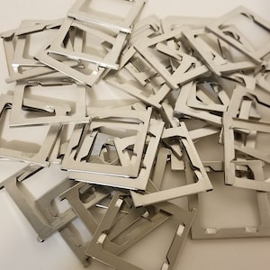 May include: A pile of small, silver-colored metal brackets. Each bracket has a rectangular frame with a series of cutouts and a bent, angled section. The brackets are scattered across a white surface, creating a dense, overlapping arrangement.