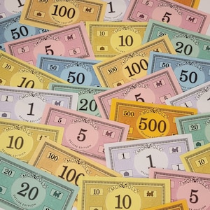 May include: A close-up of a collection of Monopoly game money in various denominations, including $1, $5, $10, $20, $50, $100, and $500. The money is printed on thick card stock and features the Monopoly logo and the words "Deluxe Edition".